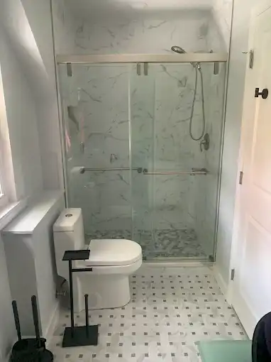 Glass shower enclosure beside a toilet in a bathroom with marble-look wall tiles.