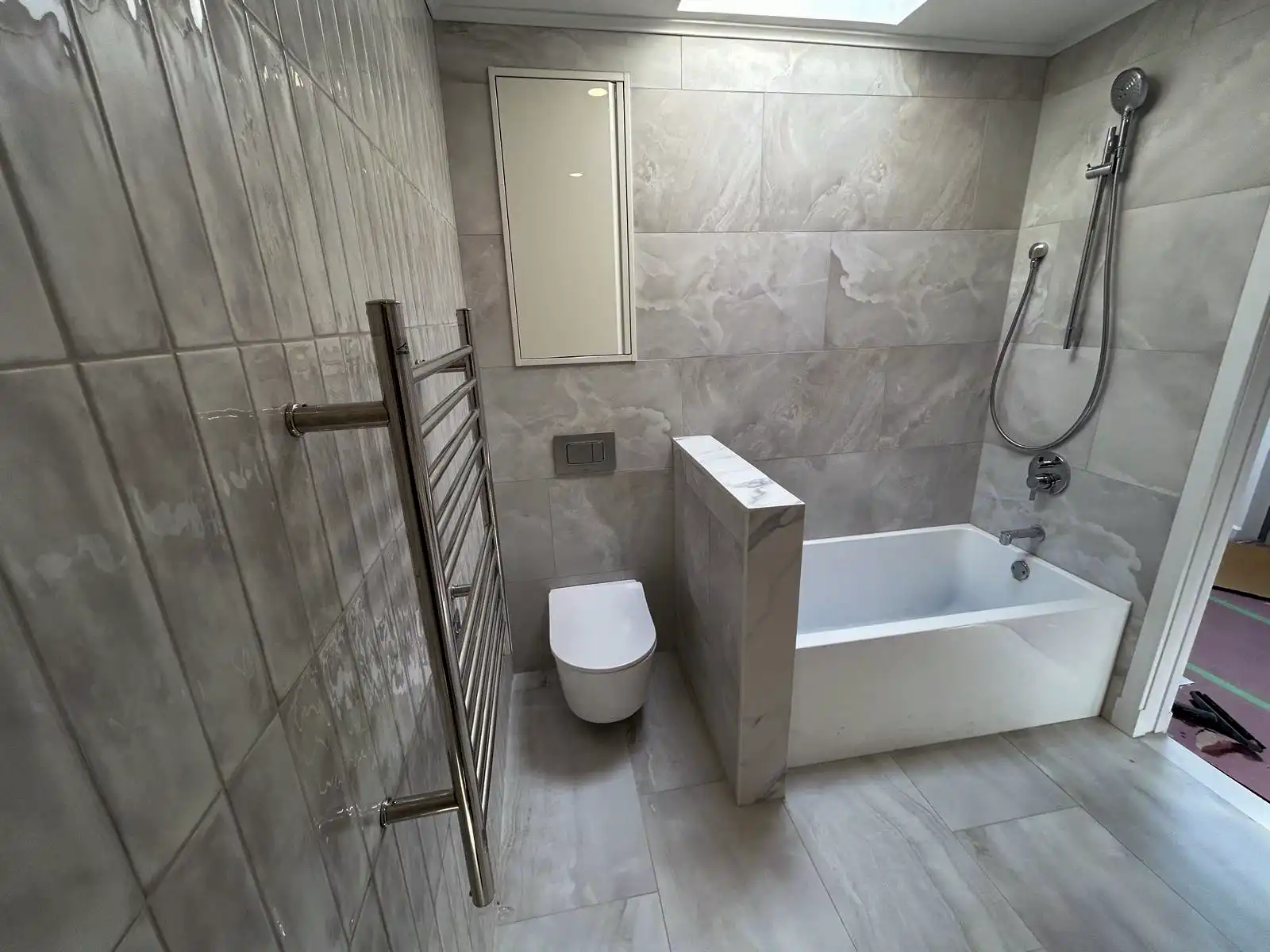 Gray-tiled bathroom with a bathtub, wall-hung toilet and chrome towel radiator.