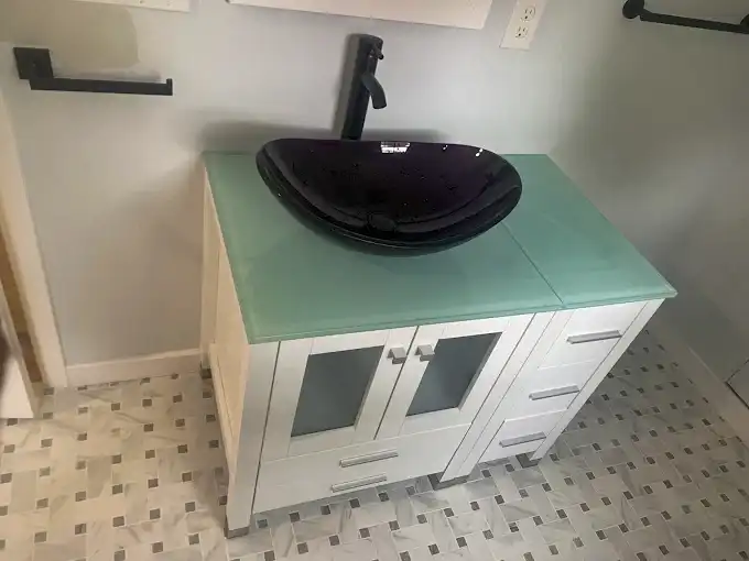 Black vessel sink on a glass countertop above a white bathroom vanity.