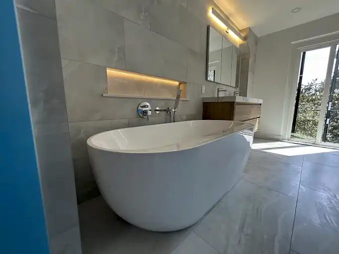Freestanding bathtub beneath an illuminated wall niche and beside a wood-finish vanity.