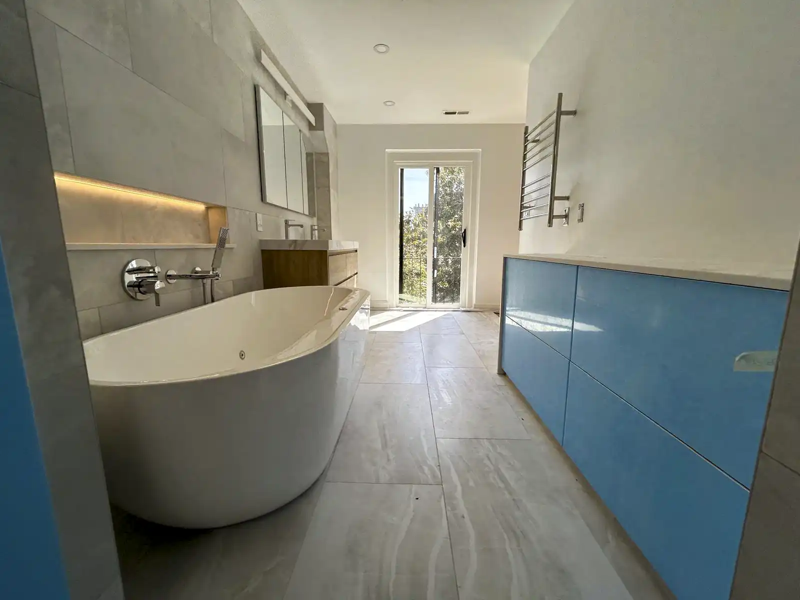 Freestanding bathtub beside tiled walls, illuminated shelving and blue cabinet fronts.